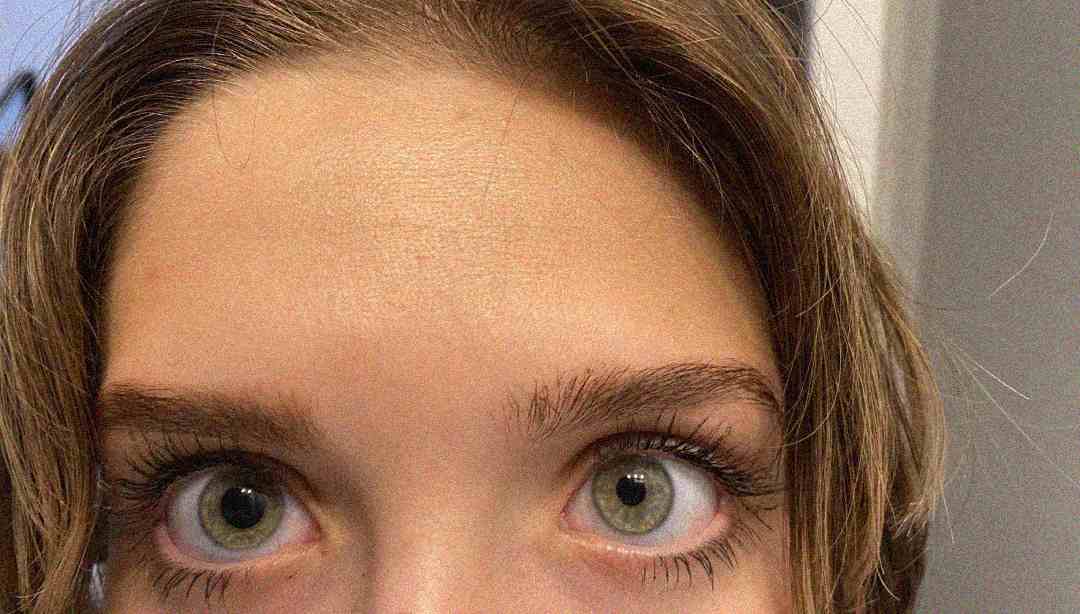 Close-up of a person's face showing wide-open green eyes, light skin, and brown hair parted in the middle. Only the upper part of the face, including eyebrows and forehead, is visible.