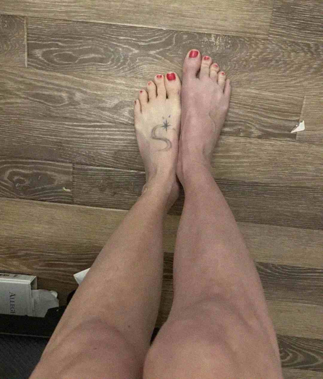 A person’s legs and feet on a wood floor, with red nail polish on the toenails and a tattoo of an "S" with a small star on the left foot.
