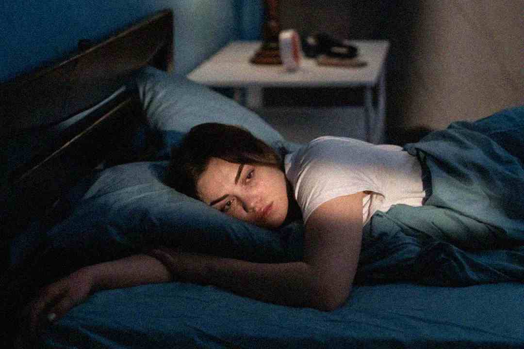 A woman lies awake in bed under blue sheets, resting on her side and looking thoughtfully towards the window. Soft morning light shines on her face, and a nightstand is visible in the background.