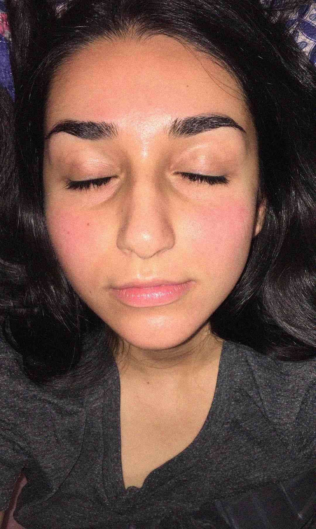 A person with long dark hair and closed eyes lies on their back, wearing a dark grey shirt. Their face appears relaxed, with slightly flushed cheeks and well-groomed eyebrows.
