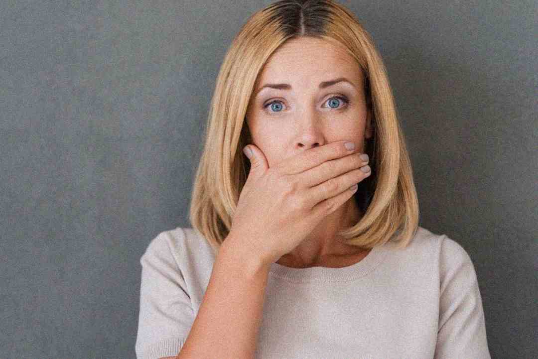 A woman with blonde hair wearing a light-colored top covers her mouth with her hand, looking surprised or shocked, against a plain gray background.