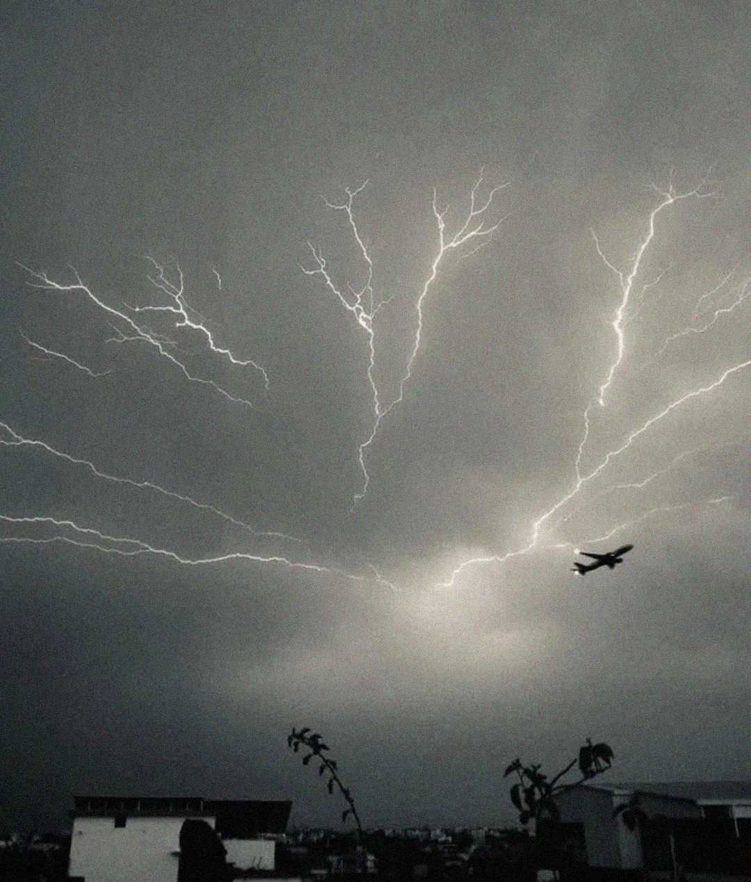 A dramatic black-and-white photo of a lightning storm at night, with several bright lightning bolts branching across the sky as a plane flies beneath them above city buildings.
