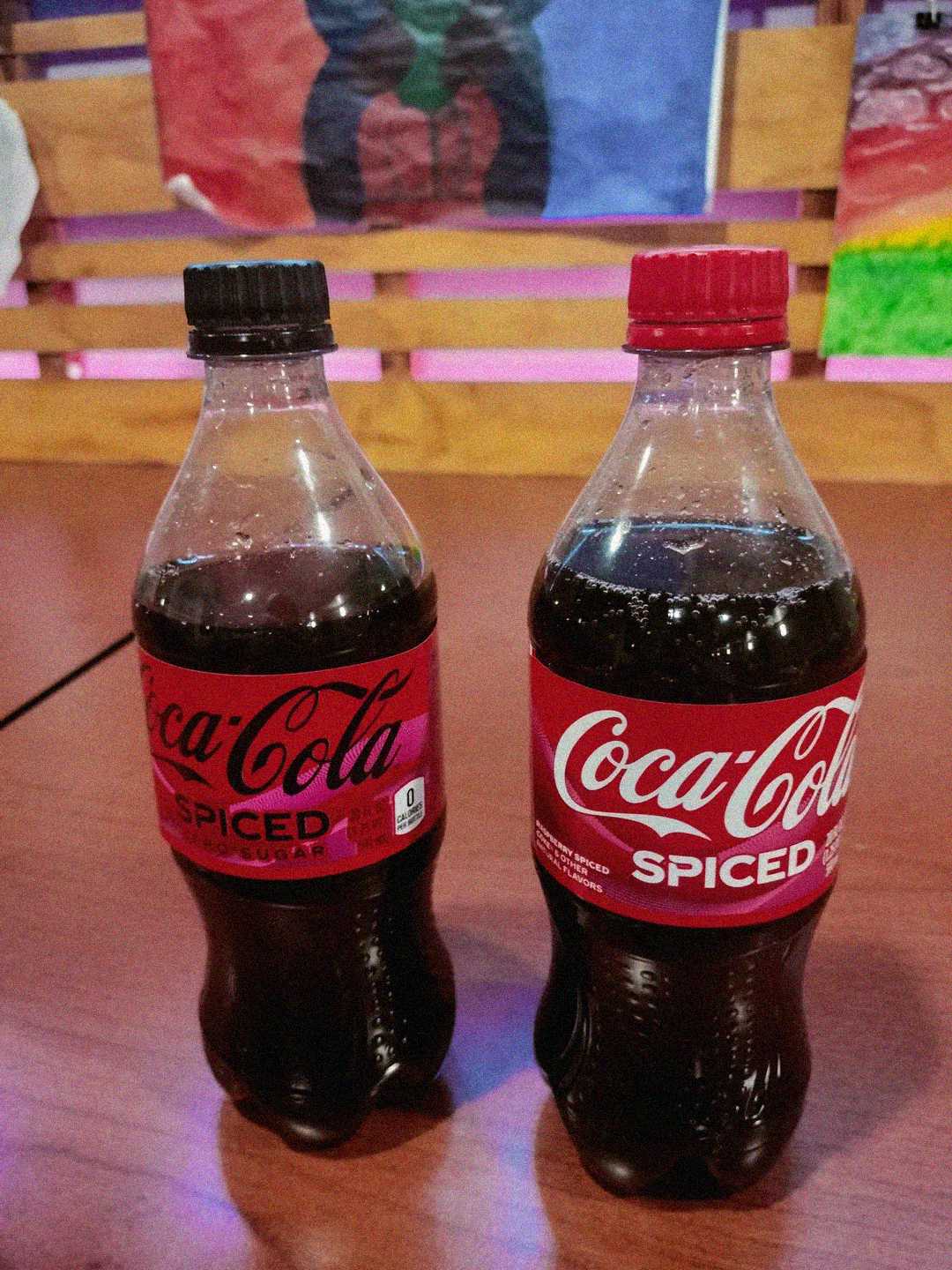 Two bottles of Coca-Cola Spiced are placed on a wooden table. The bottle on the left has a black cap and "zero sugar" label, while the one on the right has a red cap. Colorful artwork is blurred in the background.