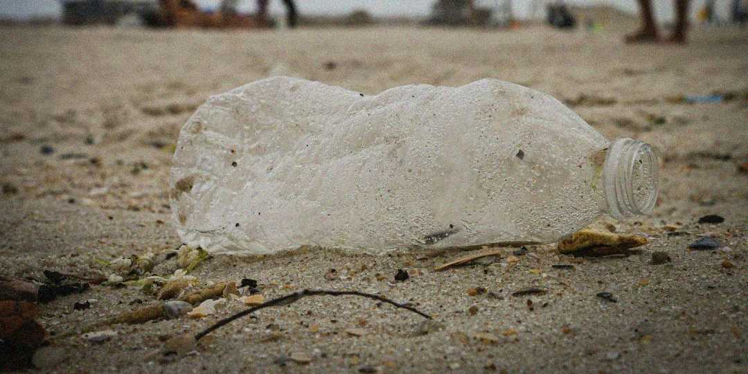 A crumpled plastic bottle lies discarded on a sandy beach, surrounded by small debris and pebbles. In the blurred background, people can be seen near the shoreline.