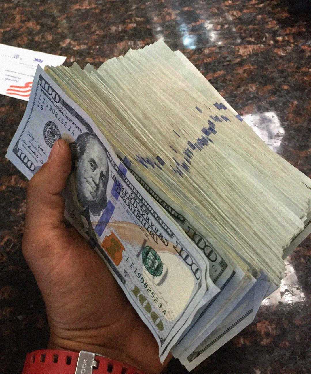 A person holding a large stack of U.S. hundred-dollar bills in their left hand over a dark marble surface.