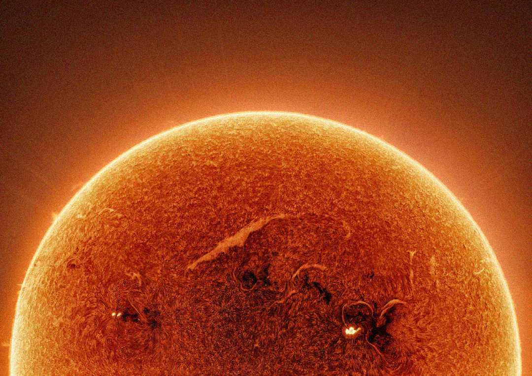 Close-up image of the Sun’s surface, showing a glowing orange sphere with visible textures, sunspots, and swirling patterns. Bright edges and solar flares create a luminous, fiery effect against the dark background.