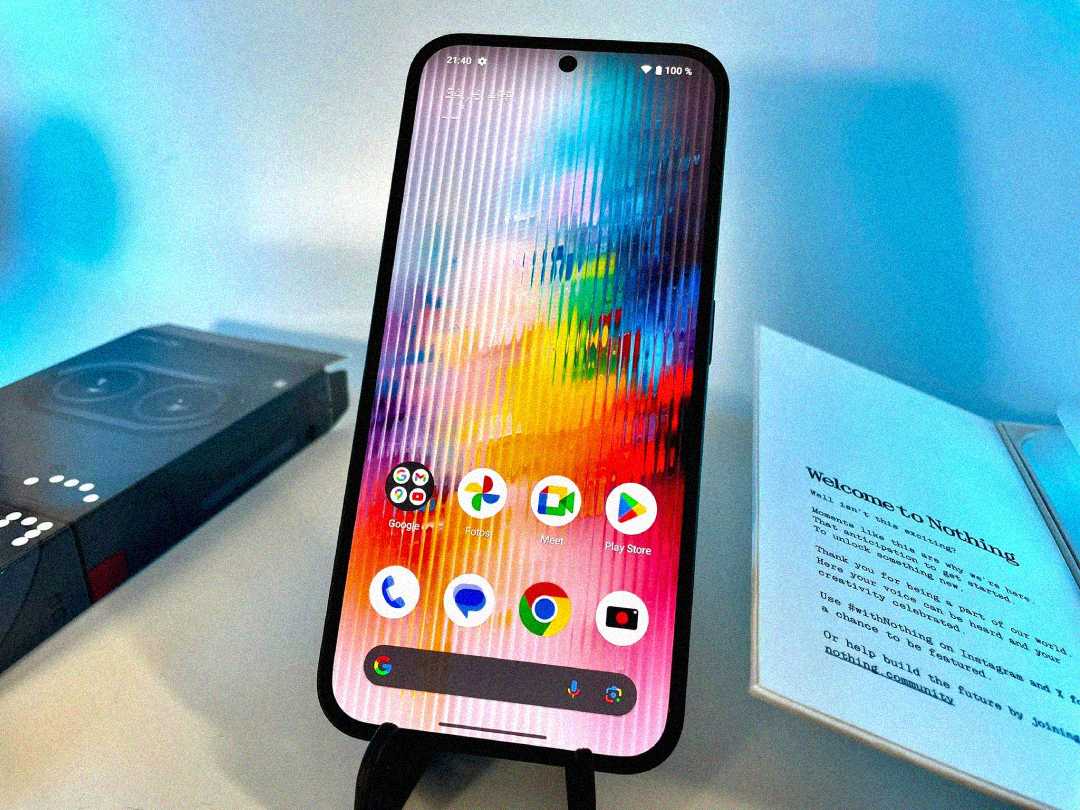 A smartphone with a colorful, glitch-like screen displays app icons on a stand. To the left is a phone box, and to the right is an open booklet that reads, "Welcome to Nothing.