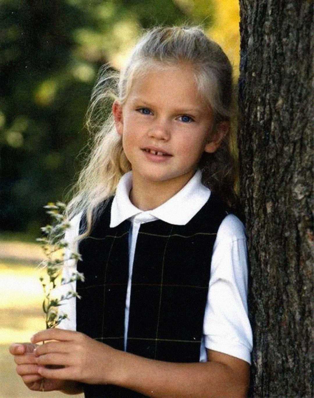 A young girl with long blonde hair, wearing a white collared shirt and black vest, stands outdoors leaning against a tree while holding a small bunch of white wildflowers. She looks at the camera and smiles slightly.