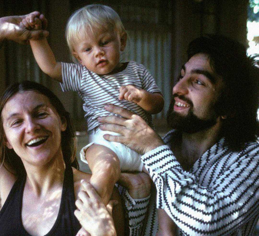 A smiling woman, a man with a beard, and a blonde toddler sit together. The man is lifting the toddler, who is holding the woman’s hand. All are wearing striped or patterned shirts.