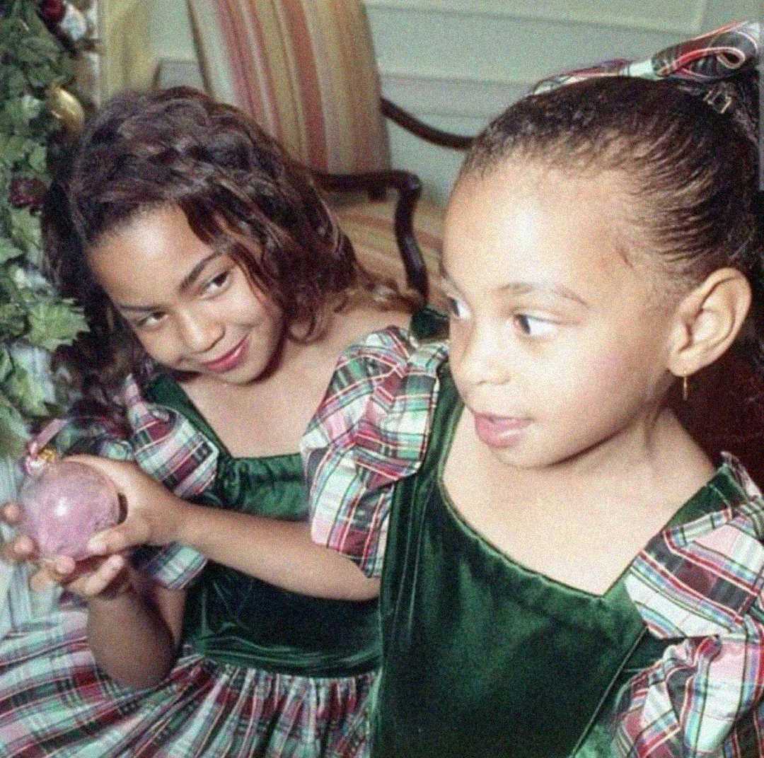 Two young girls in matching green and plaid holiday dresses sit together. One girl smiles mischievously while holding a pink ornament; the other looks off to the side, sticking her tongue out slightly.
