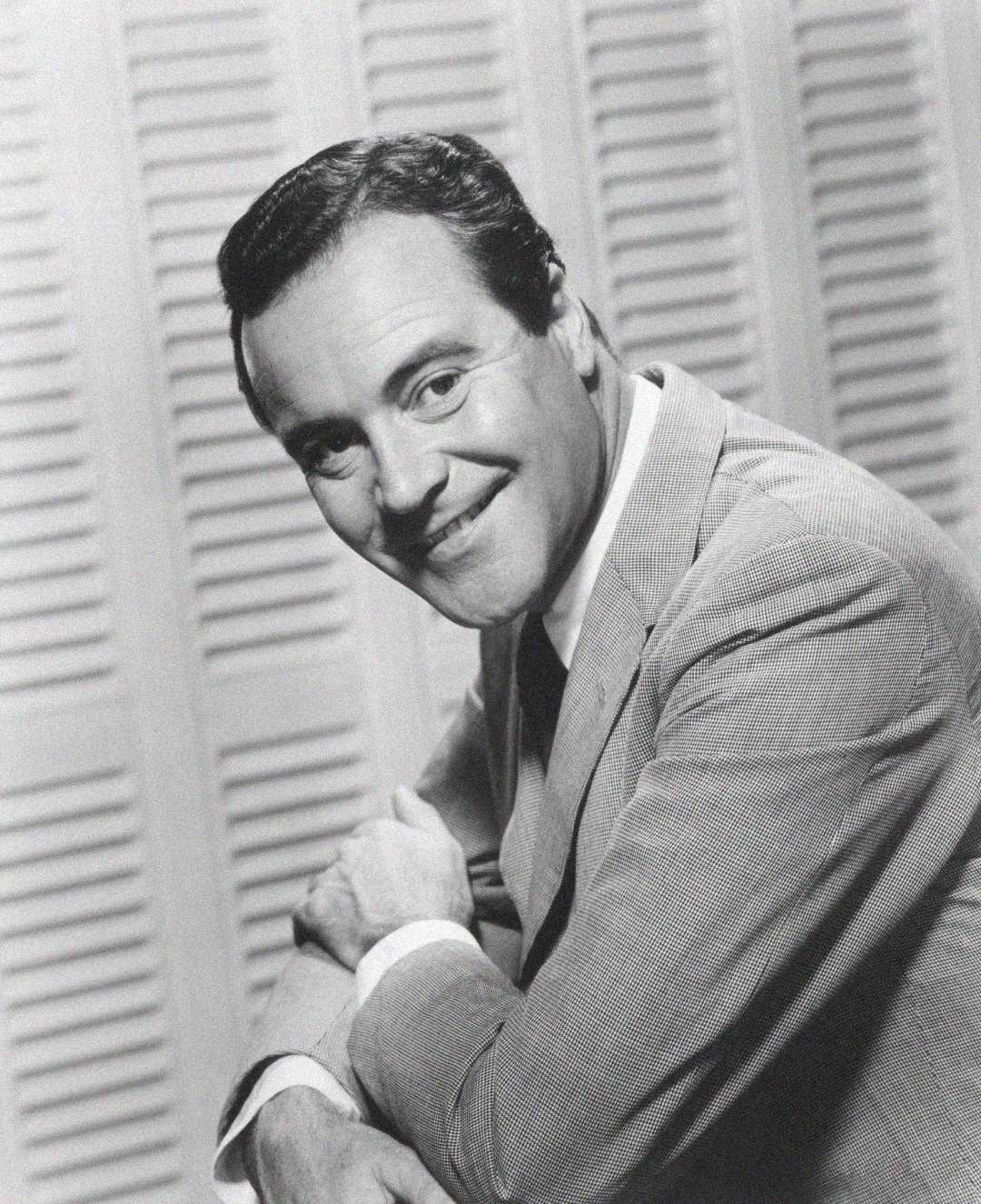 Black and white photo of a man in a light-colored suit smiling and looking at the camera, with his arms crossed and resting on a surface, in front of a backdrop with vertical slats.