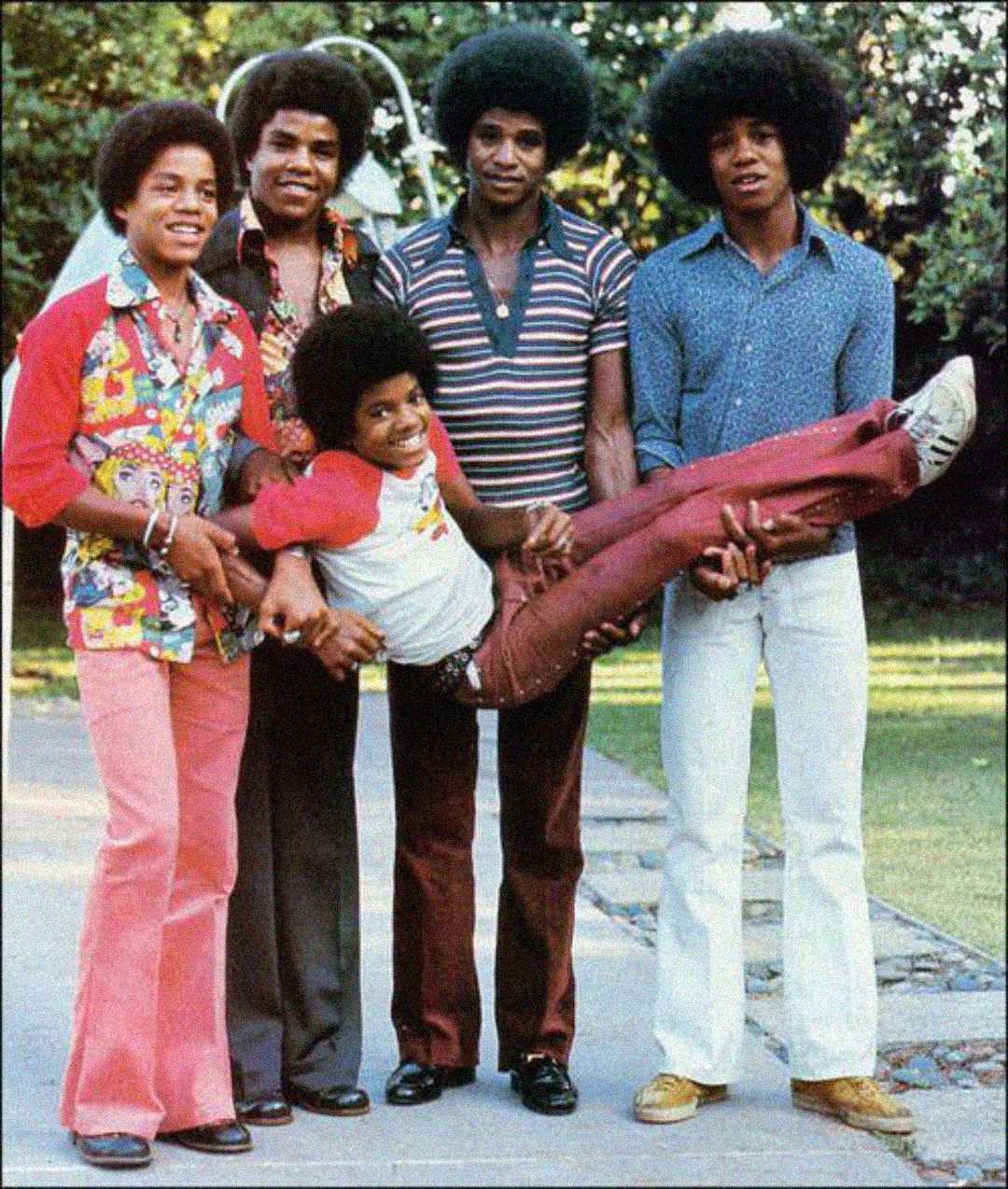 Five young men with large afros stand outdoors on a path. Four hold up the fifth, who is smiling and lying horizontally. They wear colorful, 1970s-style clothing with flared pants and patterned shirts. Trees are in the background.