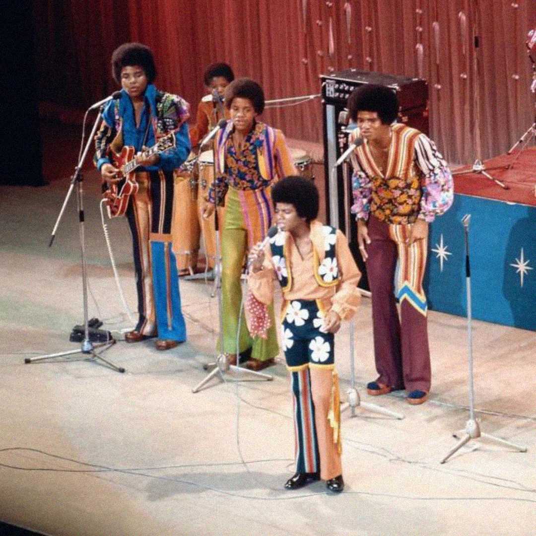 Five young male performers in colorful, patterned 1970s outfits sing and play instruments on stage with microphones, electric guitar, and drums, in front of a pink curtain backdrop.