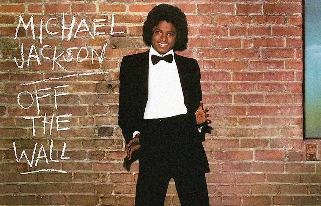 Michael Jackson in a black tuxedo with a bowtie, smiling and posing against a brick wall. "MICHAEL JACKSON OFF THE WALL" is written in white chalk-style text on the wall beside him.
