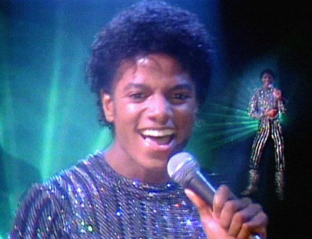 A young man wearing a sparkly shirt smiles while singing into a microphone on stage, with colorful green lights in the background and his full body visible in the distance.