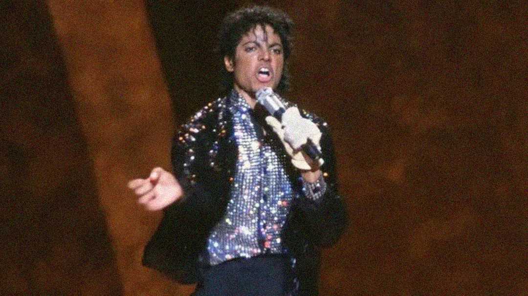 A person wearing a sequined jacket, sparkly shirt, and a white glove sings passionately into a microphone on stage, with a dramatic brown background.