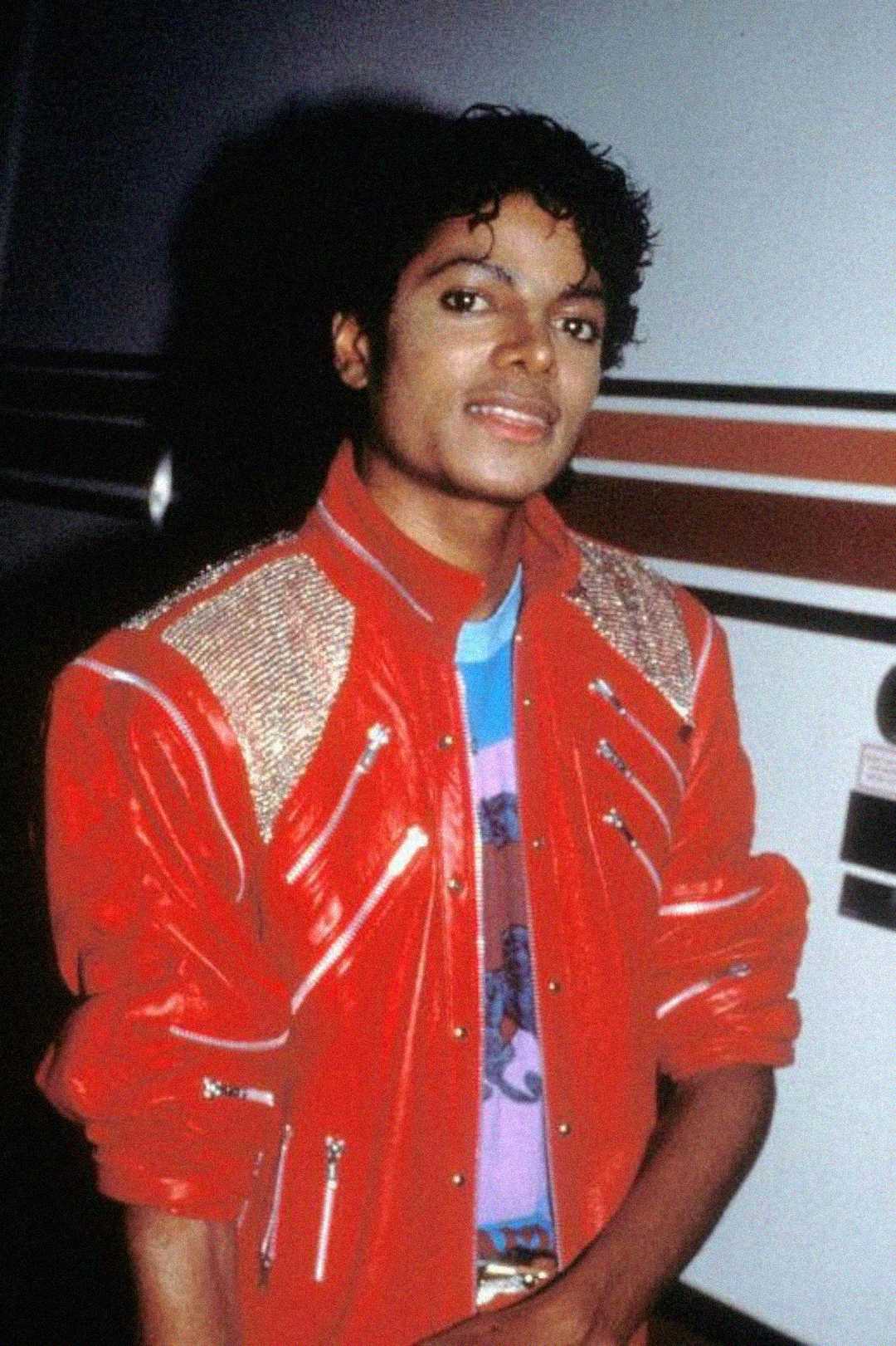 A person with curly hair wearing a shiny red jacket with metallic details and a colorful t-shirt stands in front of a wall with red and white stripes, smiling slightly at the camera.