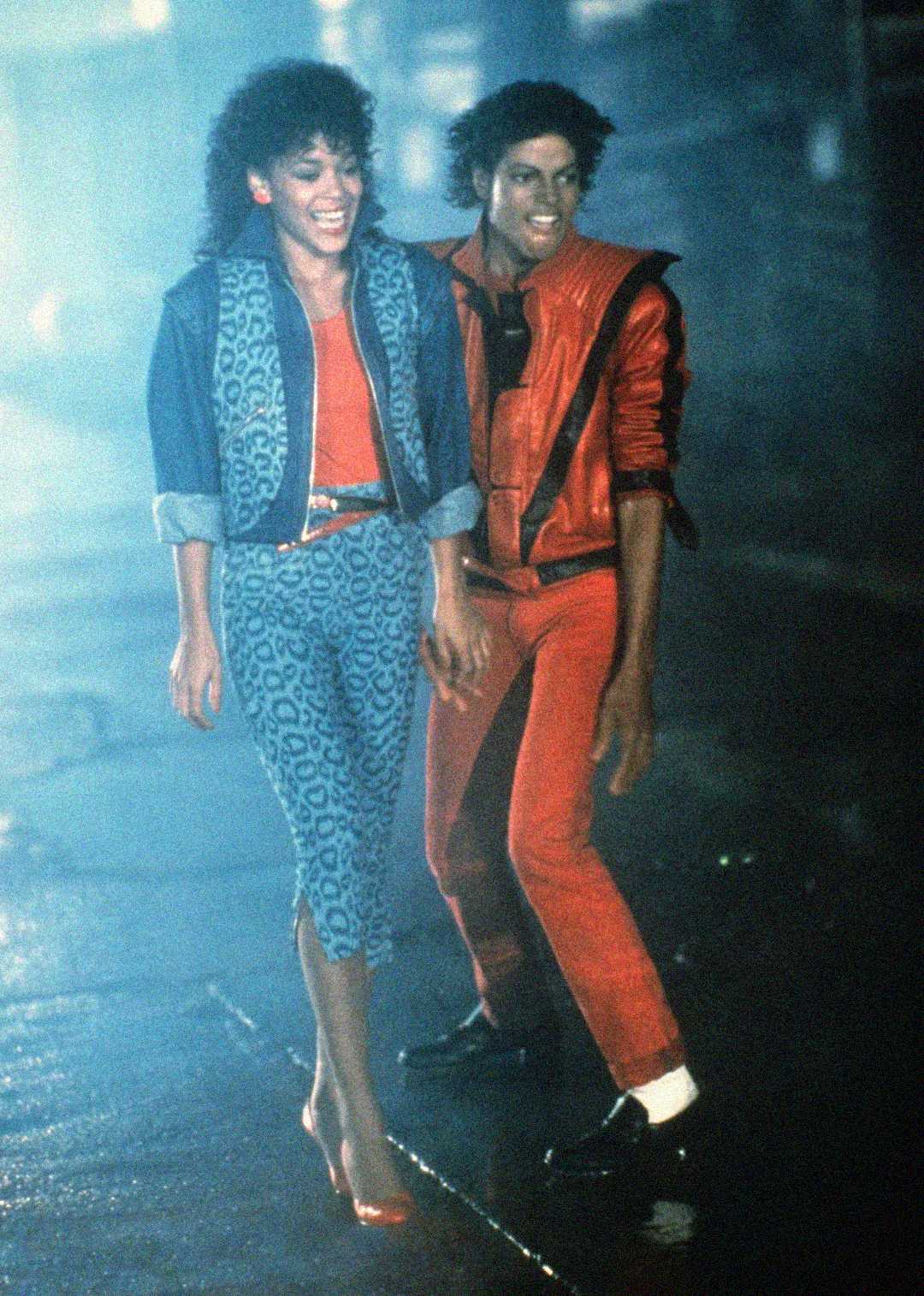 Two people in 1980s-style outfits walk down a foggy street at night, smiling and holding hands. The man wears a red jacket and pants, while the woman wears blue leopard print pants and a red top.