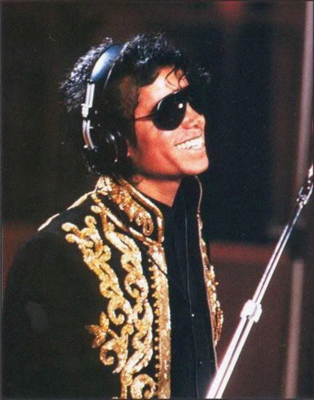 A person wearing black sunglasses, headphones, and a black jacket with ornate gold embroidery smiles while standing in front of a microphone.
