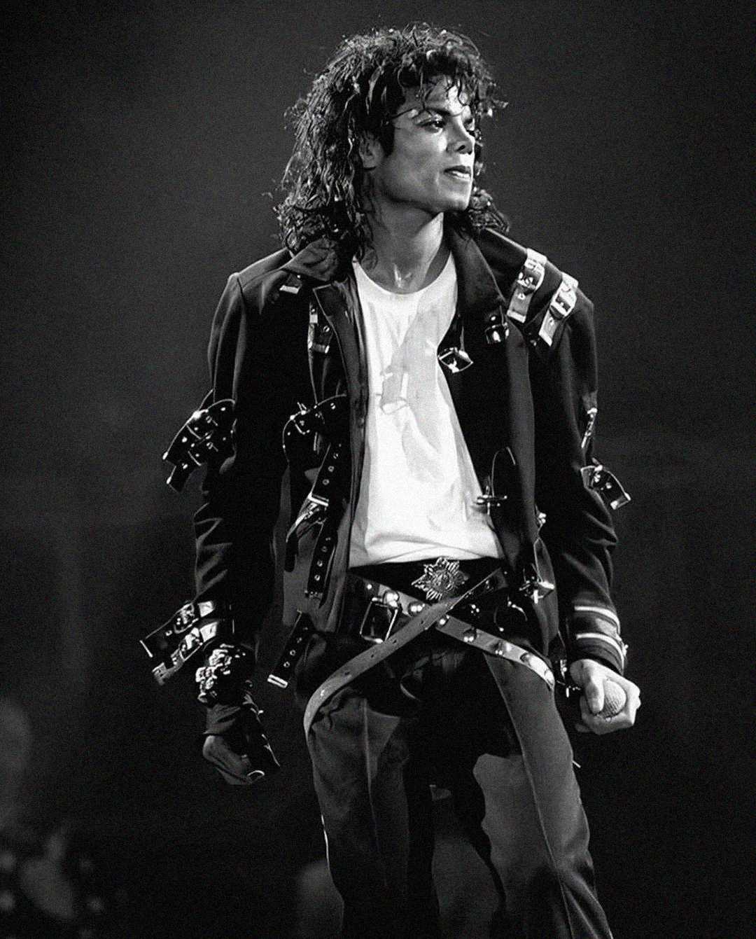 A performer with curly hair and a studded jacket stands on stage under dramatic lighting, wearing a white t-shirt, dark pants with buckles, and looking to the side with a confident expression. The image is in black and white.