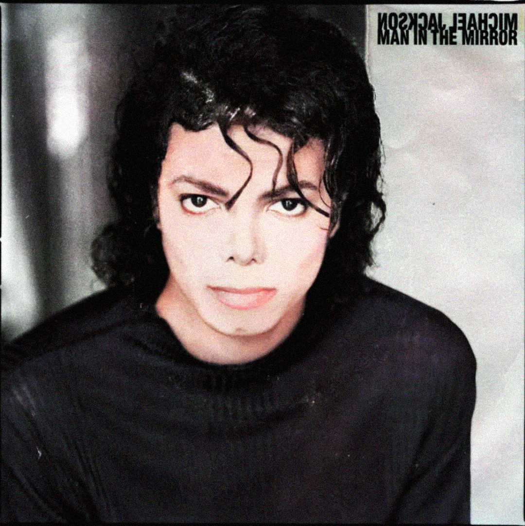 A person with curly black hair and light skin wears a black shirt, looking directly at the camera. In the upper right corner, mirrored text reads “MICHAEL JACKSON MAN IN THE MIRROR.”
