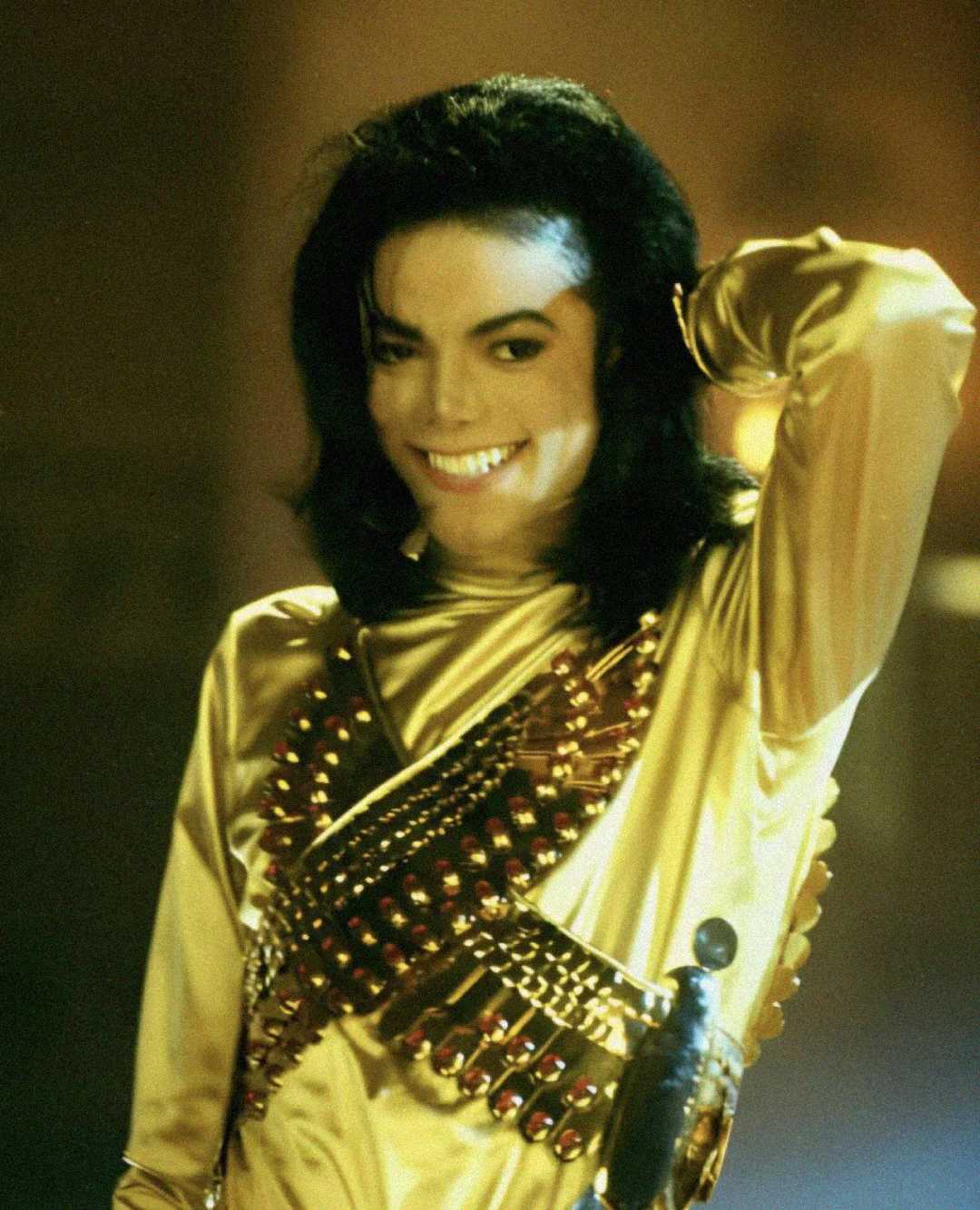 A person with long black hair wearing a shiny gold outfit with decorative details smiles and holds one hand behind their head, standing in a warmly lit setting.