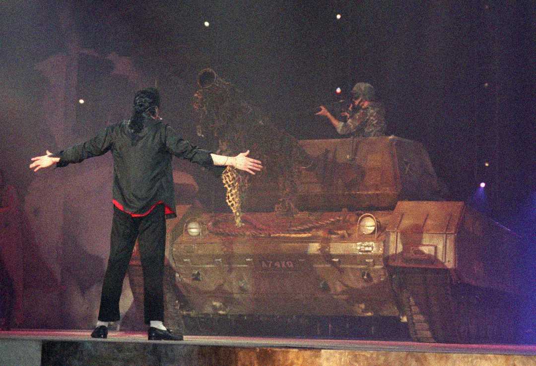 A performer in black stands with arms outstretched facing a military tank on stage, while a person in camouflage sits on top of the tank under dramatic lighting.