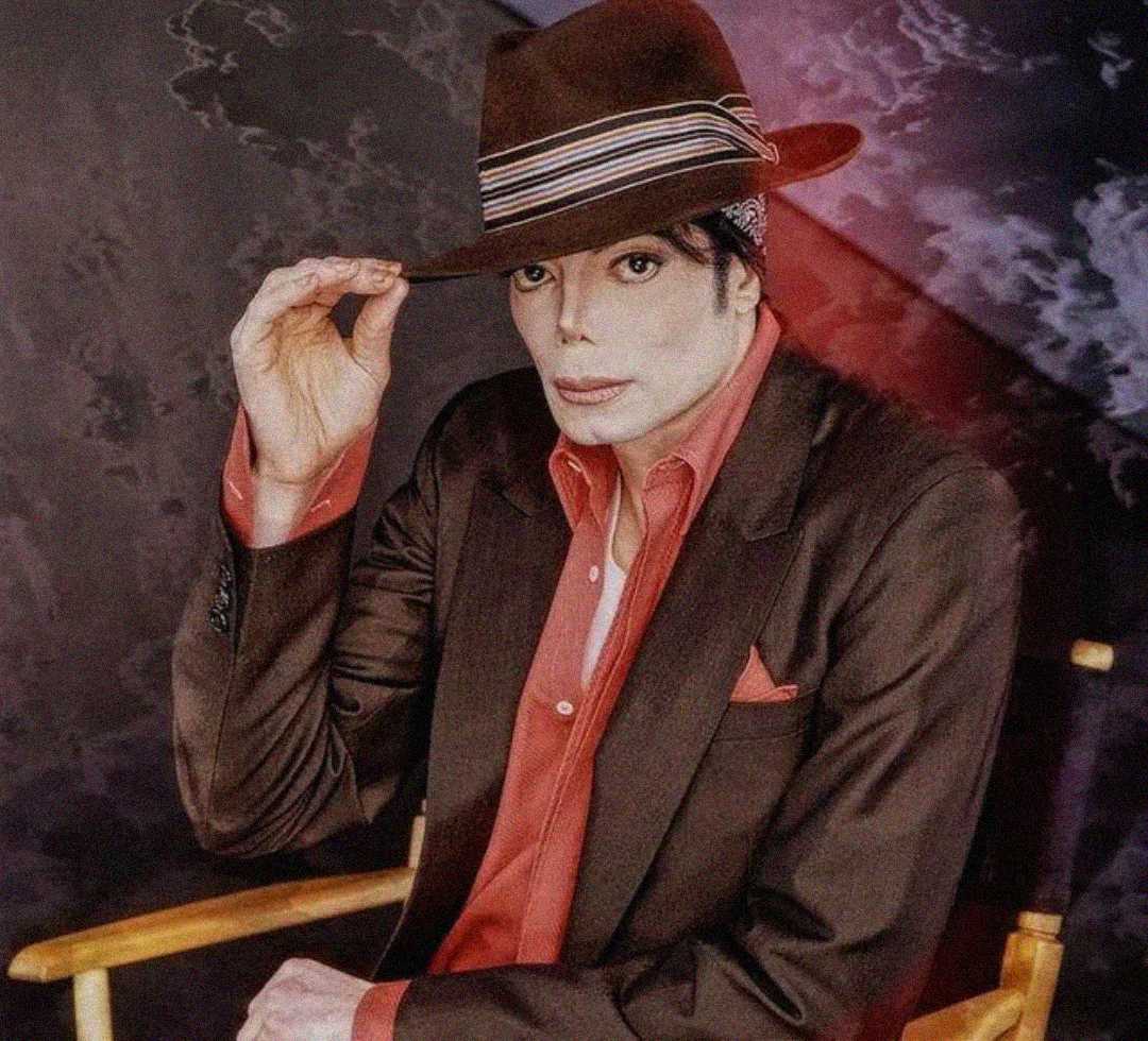 A person wearing a brown suit, red shirt, and brown fedora sits on a chair, tilting the hat with one hand. The background is dark with marbled and colored patterns.