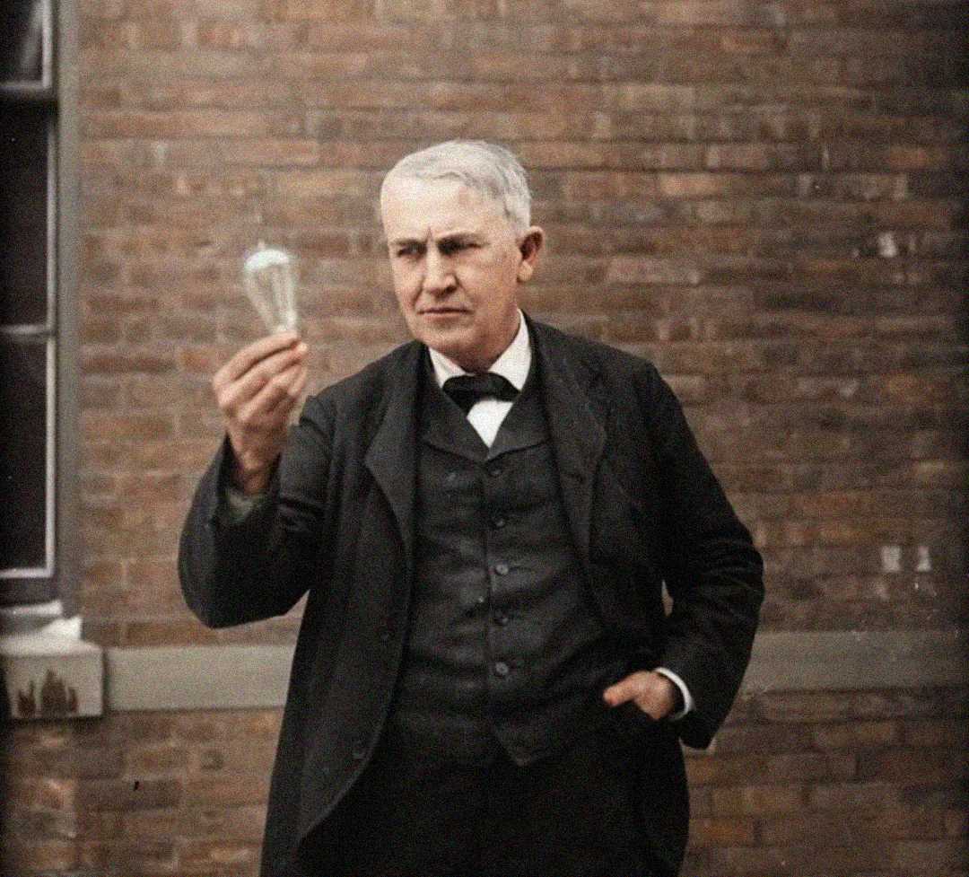 A man in a dark suit stands outside against a brick wall, holding up and examining a light bulb with a thoughtful expression.
