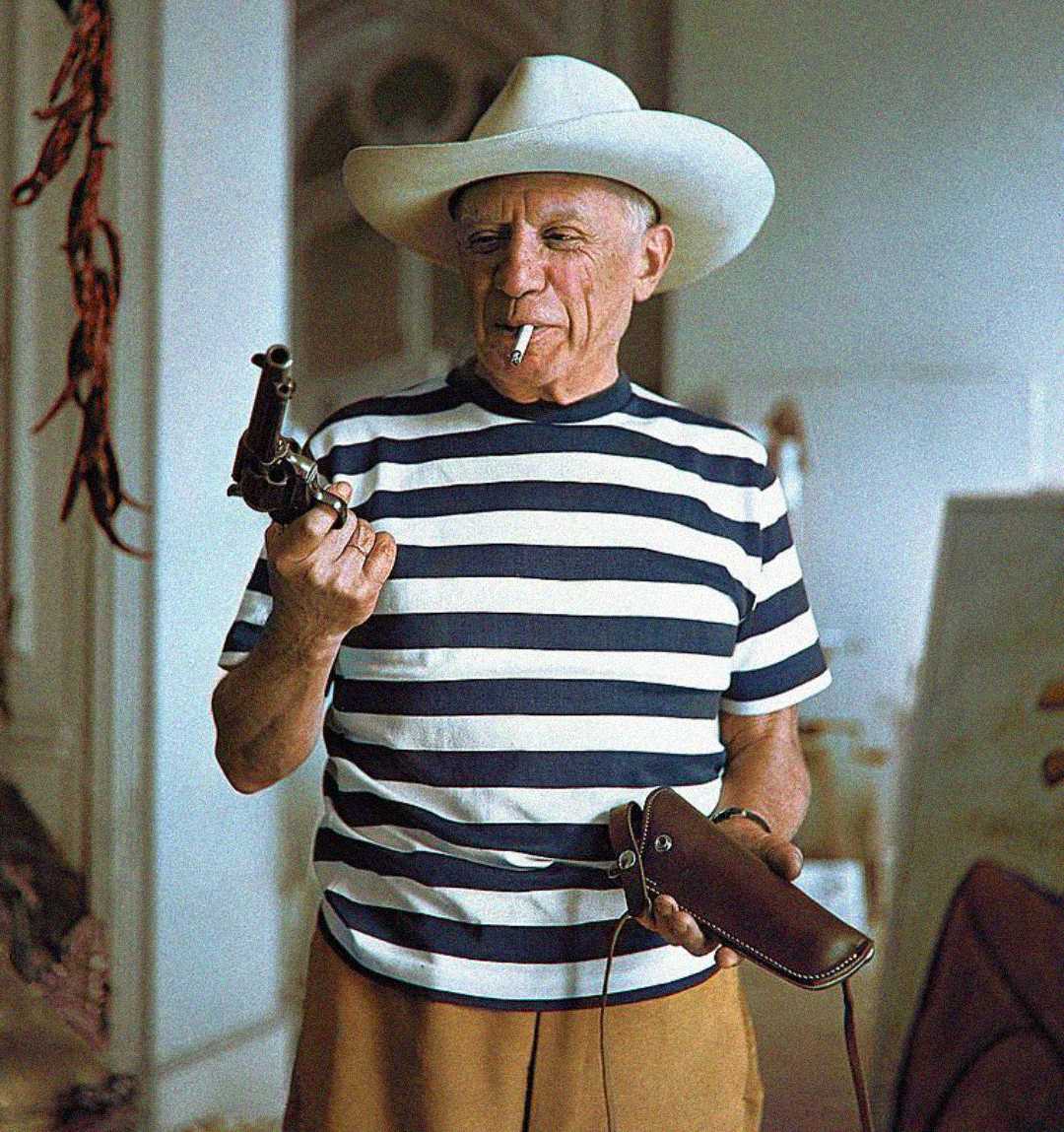 An older man in a white cowboy hat and striped shirt holds a revolver in one hand and a holster in the other, with a cigarette in his mouth, standing indoors.