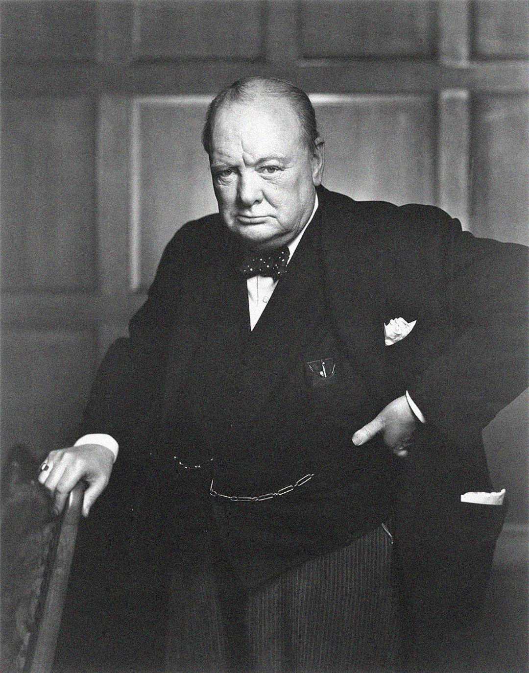 A stern-looking older man in a suit, bow tie, and pocket watch chain, stands with one hand resting on a chair and the other on his hip, against a wood-paneled background in a black and white photograph.