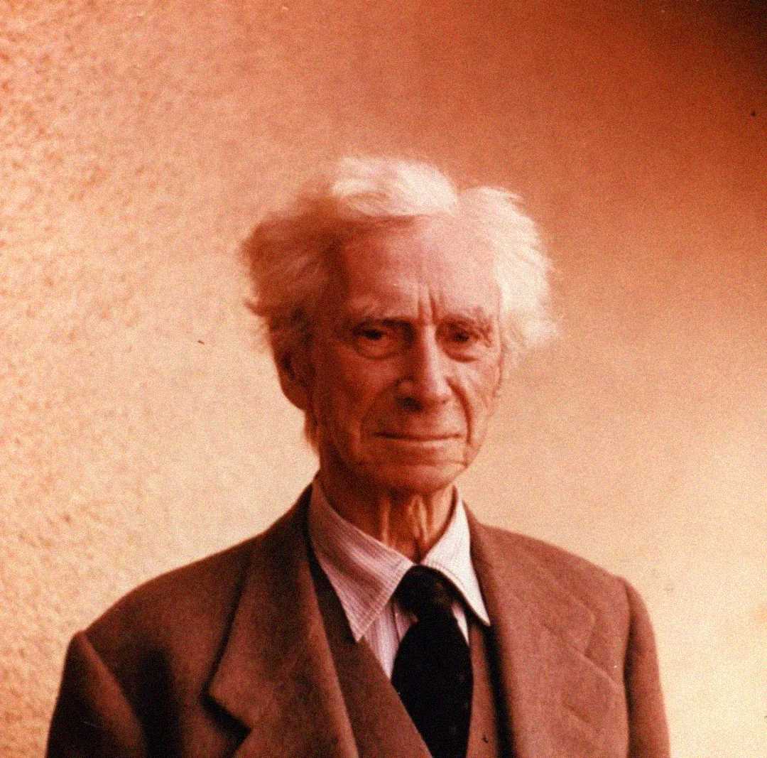 An elderly man with white hair wearing a suit, shirt, and tie stands in front of a plain, light-colored background, looking at the camera with a gentle expression.
