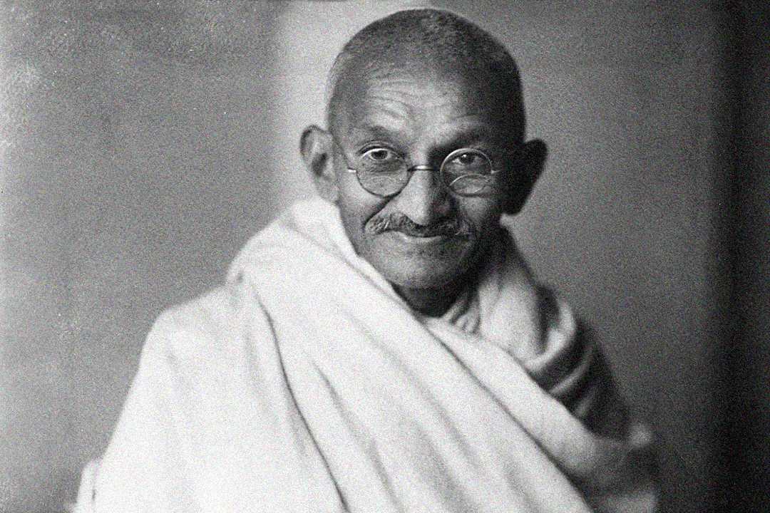 An elderly man with round glasses and a mustache smiles gently. He is wearing traditional Indian clothing, including a draped white cloth. The photo is in black and white with a soft, neutral background.