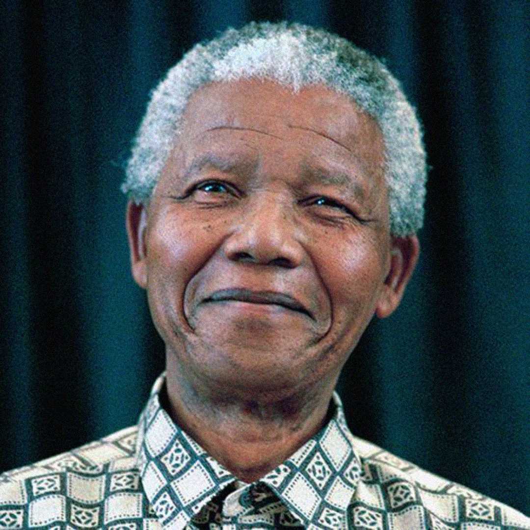 An older man with short, curly gray hair smiles gently, wearing a patterned shirt with geometric designs. The background is dark and out of focus.