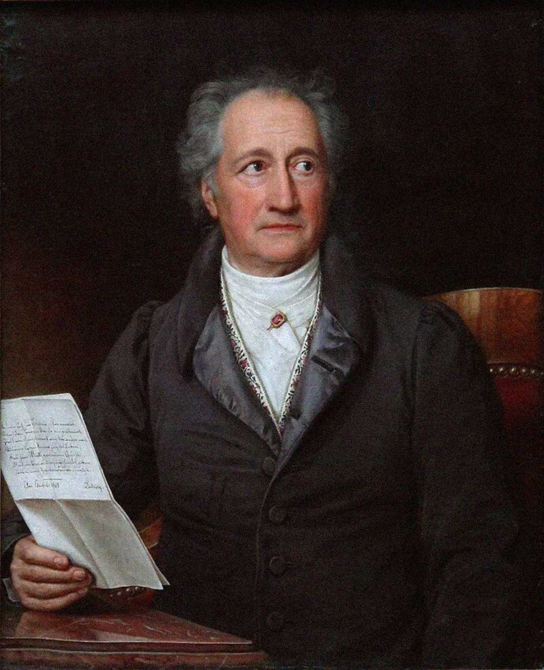 A distinguished older man with gray hair sits at a table, wearing a dark coat and white cravat. He holds a handwritten letter in his left hand and gazes thoughtfully to his right against a dark background.