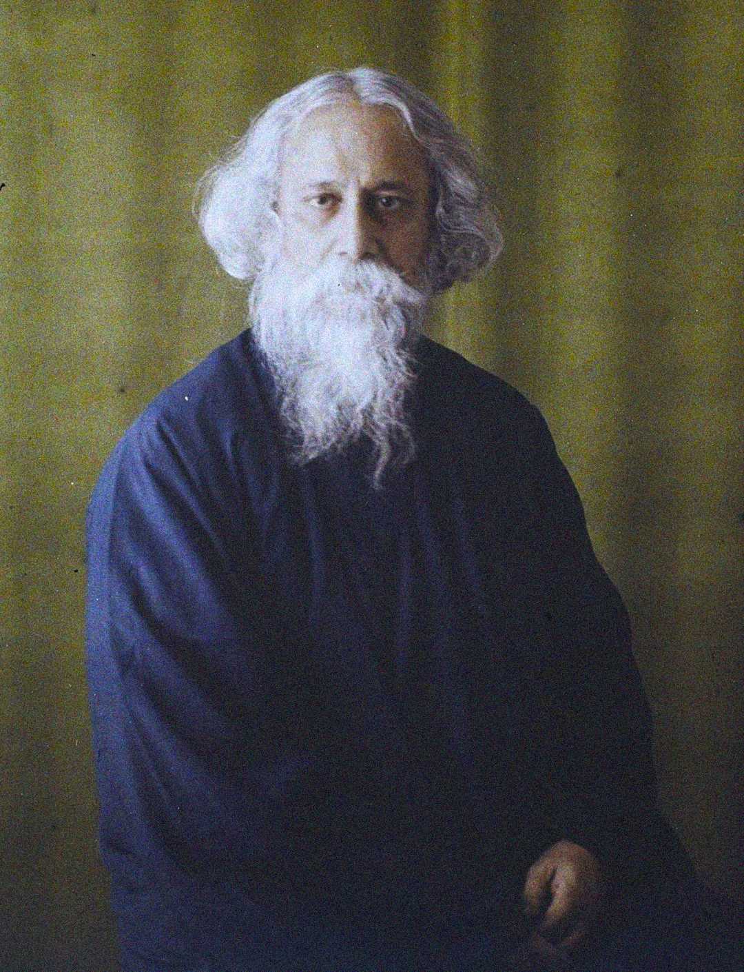 An elderly man with long white hair and a flowing white beard sits against a plain yellow-green backdrop, wearing a loose, dark blue robe, looking calmly at the camera.