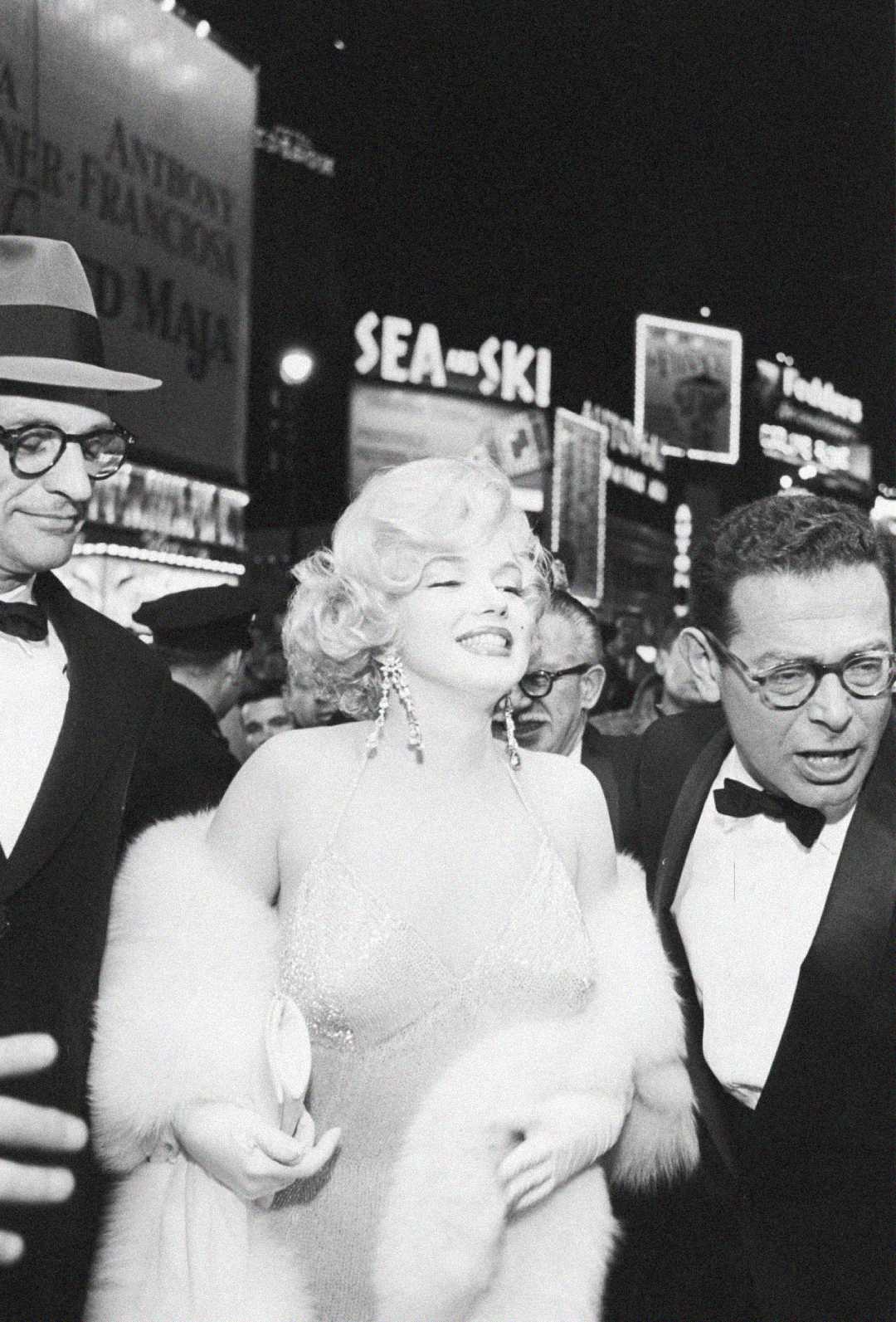 A glamorous woman in a sparkling dress and fur stole smiles amid a crowd at night in a city with bright neon signs and billboards in the background. She is flanked by two men in suits and glasses.