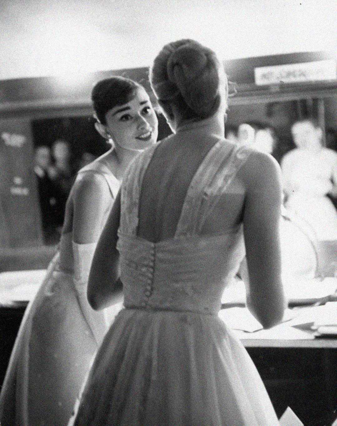 Two women in elegant, sleeveless dresses stand at a vanity in a softly lit room. One faces the mirror while the other looks over her shoulder, softly smiling. The image has a classic, vintage black-and-white style.