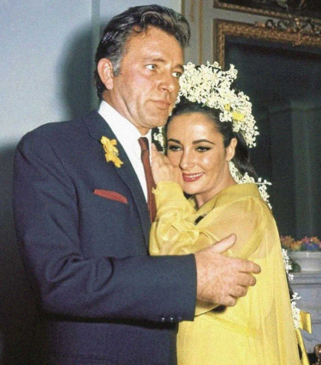 A man in a suit and a woman in a yellow dress with a white floral headpiece embrace and smile. The woman rests her head on the man's chest in a warmly lit, elegant room.