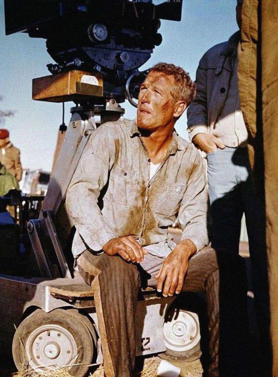 A man with dirt on his face and clothes sits on a piece of film equipment outdoors, looking up. He wears a light, worn shirt and is surrounded by crew members; a large movie camera is behind him.