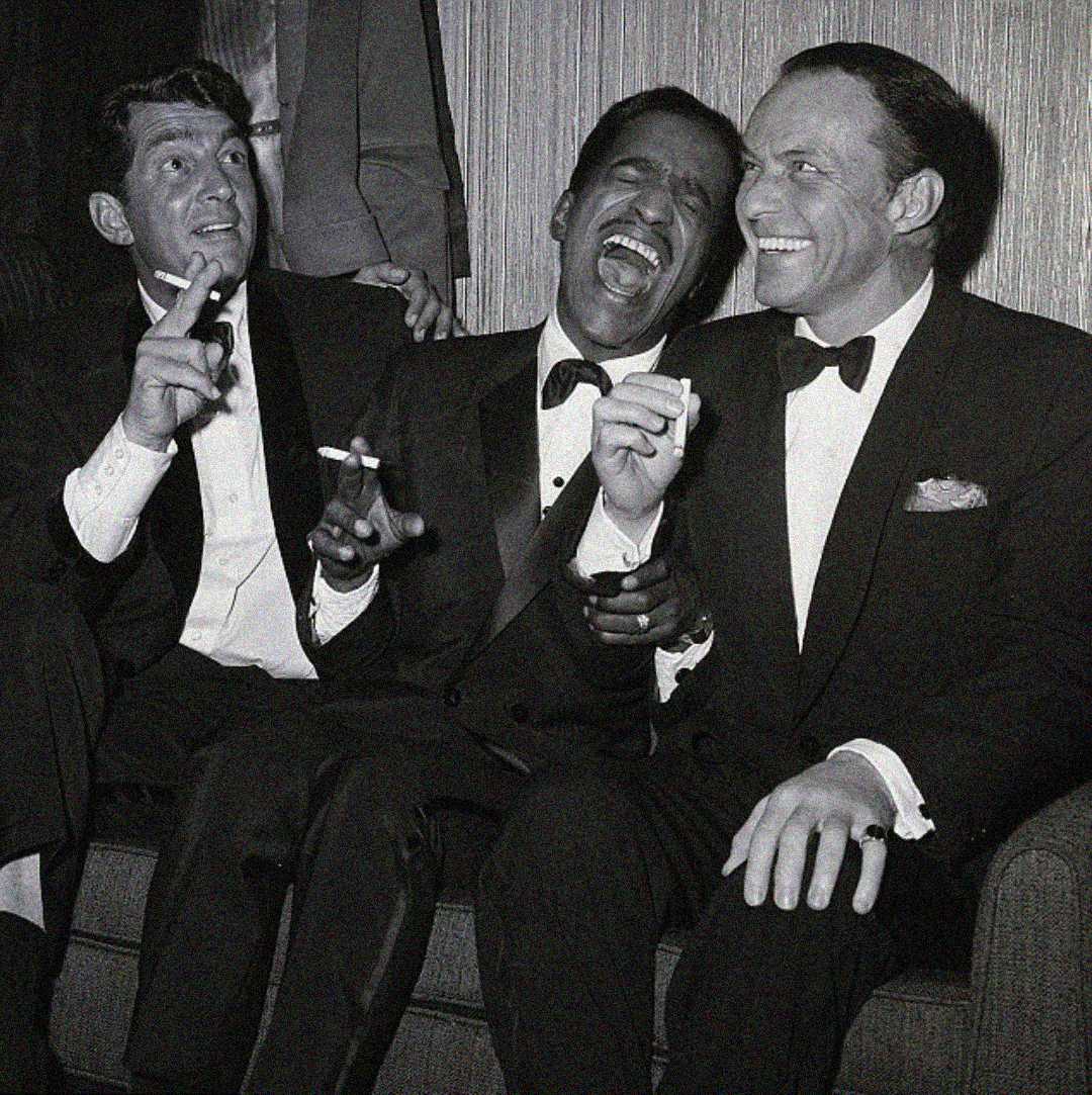 Three men in tuxedos sit together on a couch, holding cigarettes and laughing joyfully. The image is in black and white, suggesting it’s from a past era, and the mood is lighthearted and celebratory.