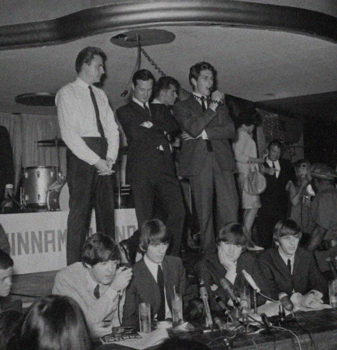 A black-and-white photo shows the Beatles seated at a table with microphones during a press conference. Several men in suits stand behind them on a stage, and other people are visible in the background.