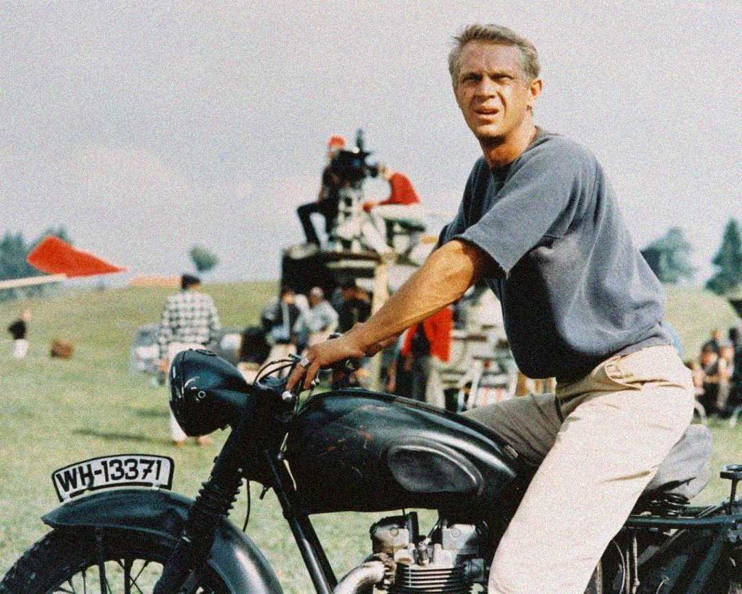 A man in a grey shirt and light pants sits on a classic black motorcycle outdoors, with people and vehicles in the background on a grassy field.