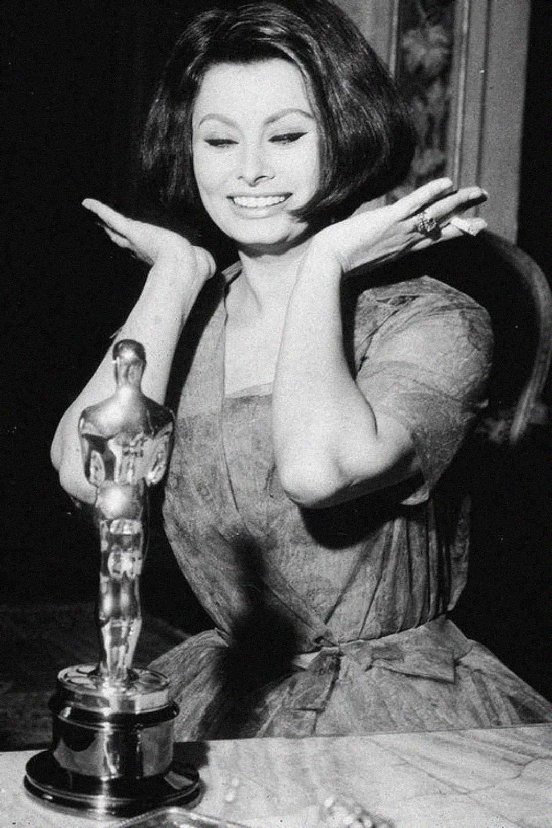 A woman in a vintage dress smiles joyfully with her hands raised near her face while seated in front of an Oscar statuette on a table. The image is in black and white.
