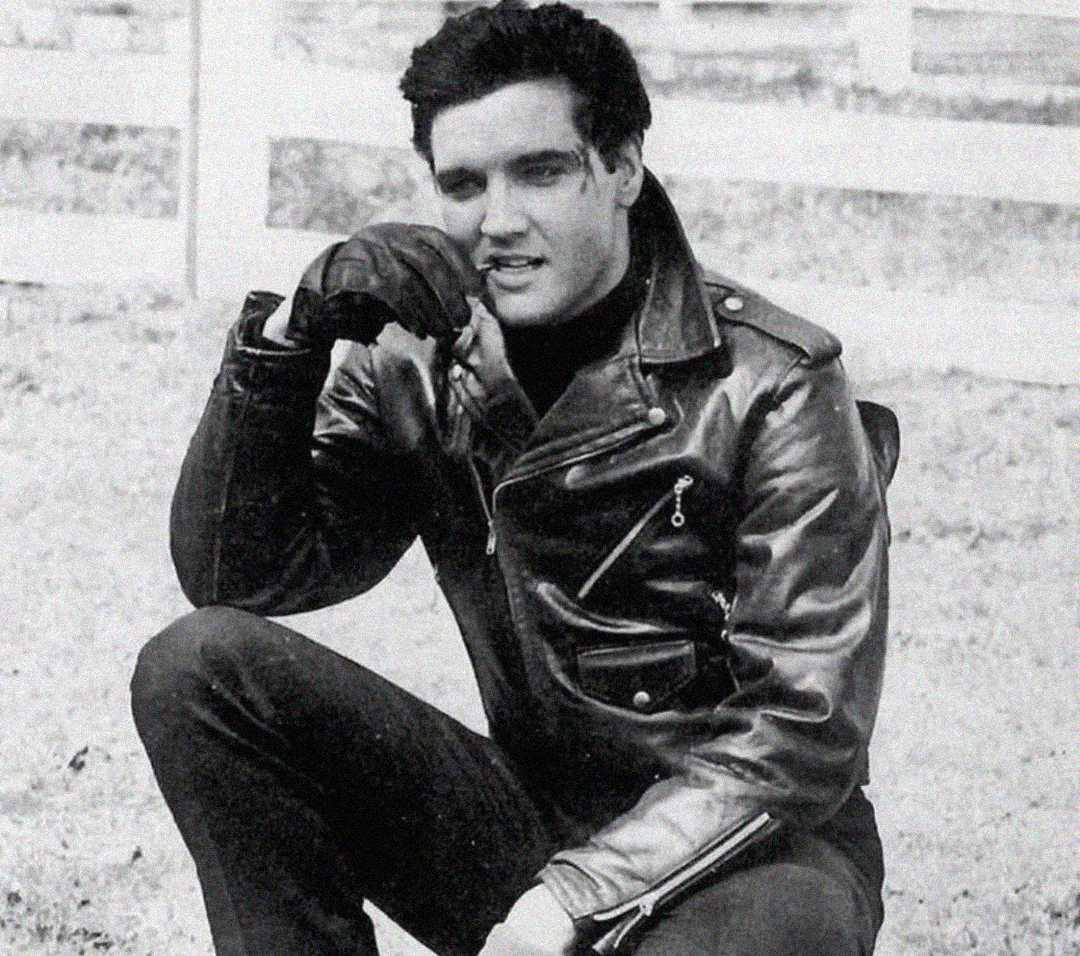 A man in a black leather jacket and gloves crouches outdoors, resting his elbow on his knee and his hand near his mouth, with a white fence and grass in the background. The image is black and white.