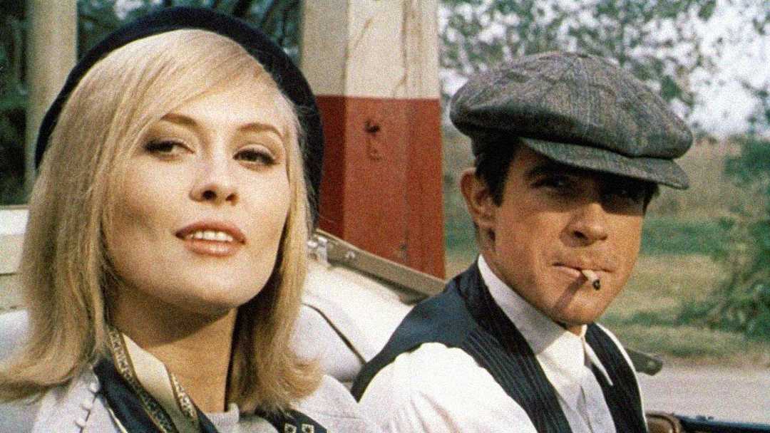 A woman with blonde hair and a black beret sits beside a man wearing a flat cap and vest, who has a cigarette in his mouth. They are in an old-fashioned car with greenery in the background.