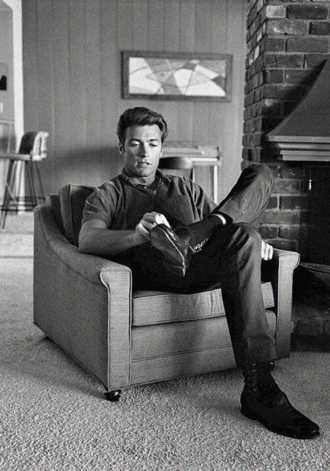 A man sits in a cushioned armchair, one leg crossed over the other, adjusting his shoe. He wears a short-sleeved shirt and dark pants in a mid-century styled living room with a brick fireplace. The photo is black and white.