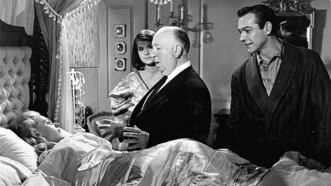 A black-and-white scene shows three people standing around a woman lying in bed, covered by a blanket. The setting appears to be a luxurious bedroom with ornate decor.