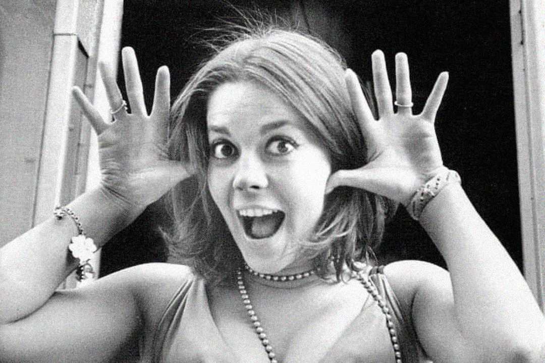 A young woman with shoulder-length hair smiles widely and holds her hands up by her face, fingers spread, giving a playful expression. She wears bracelets, rings, and a beaded necklace. The image is in black and white.