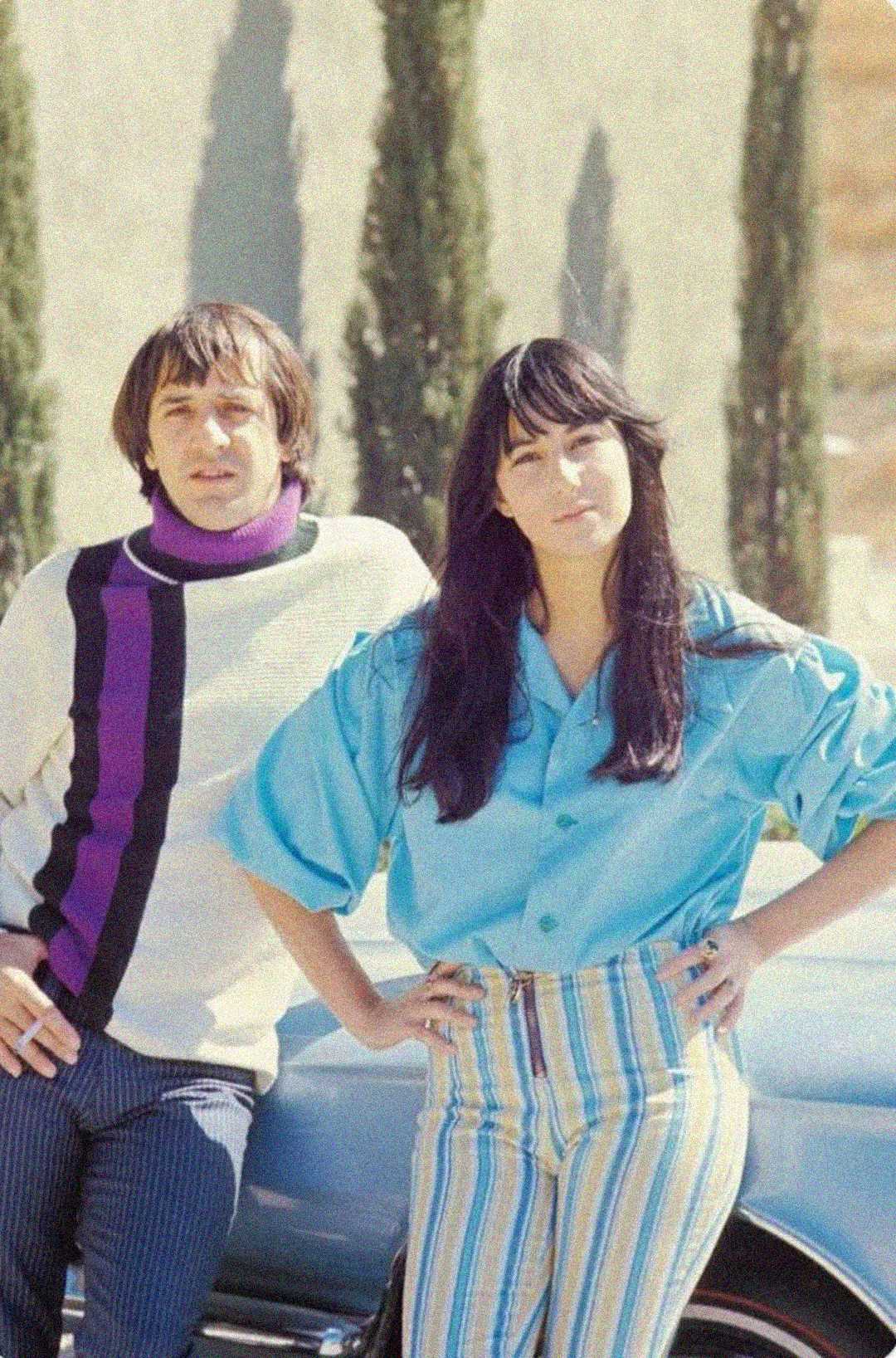 A man in a white sweater with a purple and black stripe and a woman in a bright blue blouse with striped pants stand outdoors by a car, with tall trees and a light wall in the background.